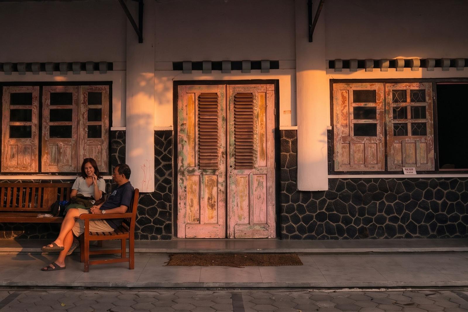 The front of Monti Kopi, with classic teak doors and two guests on a wooden bench.