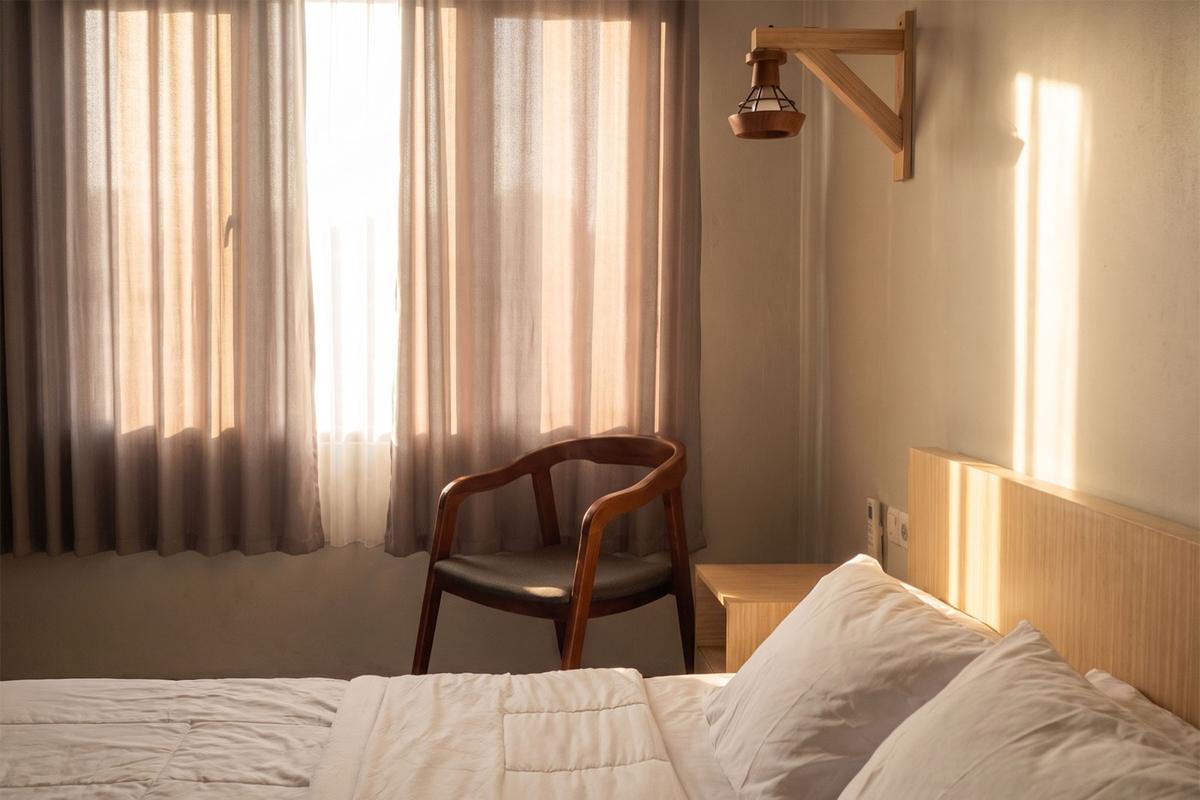 Double bed and wooden chair by curtained windows with soft morning light.